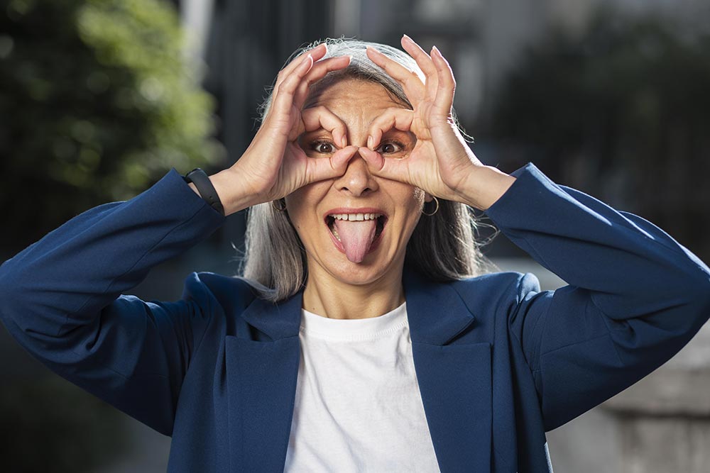 Femme entre cinquante et soixante ans souriante aux cheveux gris, faisant une grimace joyeuse avec ses mains en lunettes autour des yeux. Son attitude positive incarne l' état d'esprit positif et résilient indispensable face aux aléas de la vie.