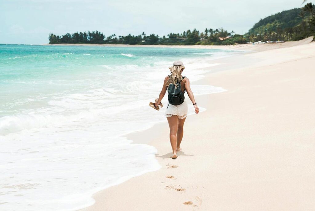 Une femme, vue de dos, marche paisiblement le long d'une magnifique plage, à la limite du bord de l'eau. Elle porte un short et un tee-shirt, tenant ses tongs à la main. Cette photo illustre les bienfaits de la marche à pied, mettant en lumière le calme et la détente qu'offre cette activité.