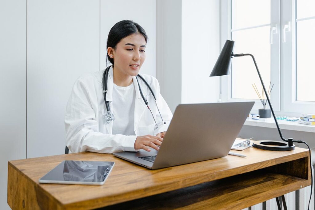 Une femme médecin, portant une blouse blanche et un stéthoscope autour du cou, est assise à son bureau dans son cabinet. Elle consulte son ordinateur, suggérant qu'elle examine des données médicales ou prend des notes.