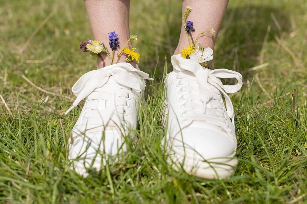 Gros plan sur une paire de chaussures de sport blanches portées par une femme, visible des pieds jusqu'aux chevilles, dans un environnement naturel. De petites fleurs champêtres décorent les chaussures, apportant une touche de fraîcheur et de joie.