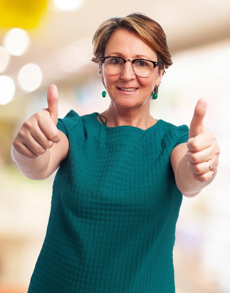 Femme d'environ cinquante ans, portant des lunettes et une robe verte, affiche un sourire satisfait tout en levant les deux pouces vers l'avant en signe d'approbation.