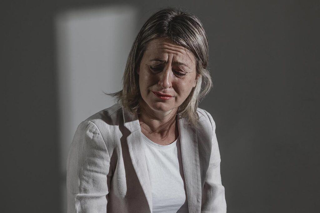 Femme d'environ cinquante ans, sur un fond sombre et gris, les larmes aux yeux, le regard baissé vers le sol.