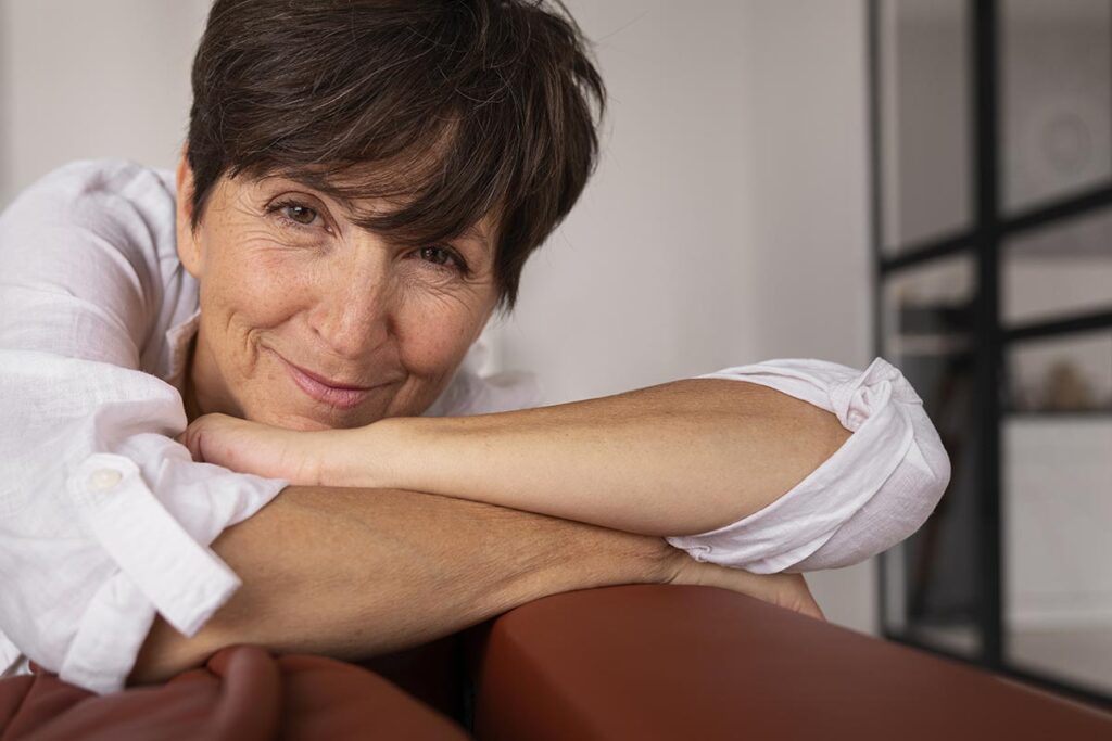 Gros plan d'une femme souriante dans la cinquantaine, la tête reposant doucement sur ses bras croisés sur le dossier de son canapé.