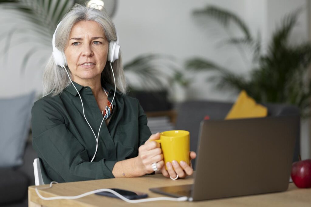 Femme entre quarante et cinquante ans, vêtue d'un chemisier gris foncé, aux cheveux longs grisonnants, assise à un bureau devant son ordinateur. Elle porte un casque audio et tient une tasse de thé dans les mains, visiblement concentrée sur ce qu'elle écoute. Des plantes légèrement floues en arrière-plan suggèrent qu'elle est dans son domicile.