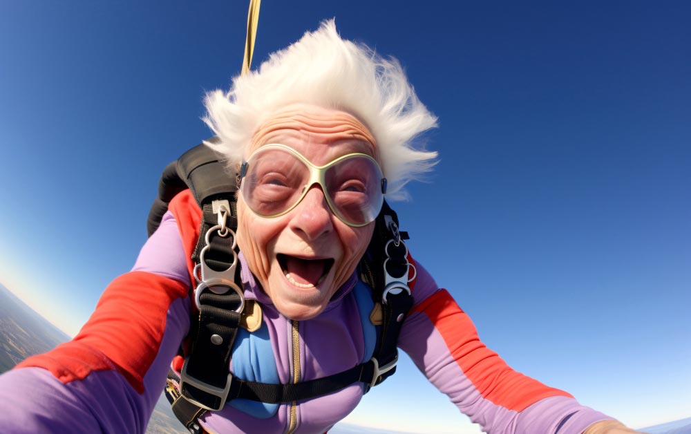 Une mamie aux cheveux blancs de plus de soixante-dix ans saute en parachute, revêtue d'une combinaison de parachutiste violette avec des bandes rouges. Son visage est ébouriffé par la descente en chute libre avant l'ouverture du parachute. Malgré l'adrénaline, son sourire radieux exprime une joie immense de réaliser son rêve. Cette image prouve qu'il n'y a pas d'âge pour concrétiser ses aspirations.