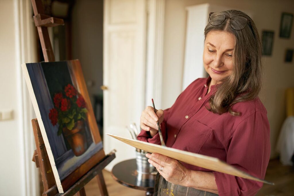 Femme dans la cinquantaine se tenant devant un chevalet, sur lequel est posée une toile représentant un vase avec des fleurs rouges. Elle mélange soigneusement des couleurs sur sa palette, prête à poursuivre sa peinture.