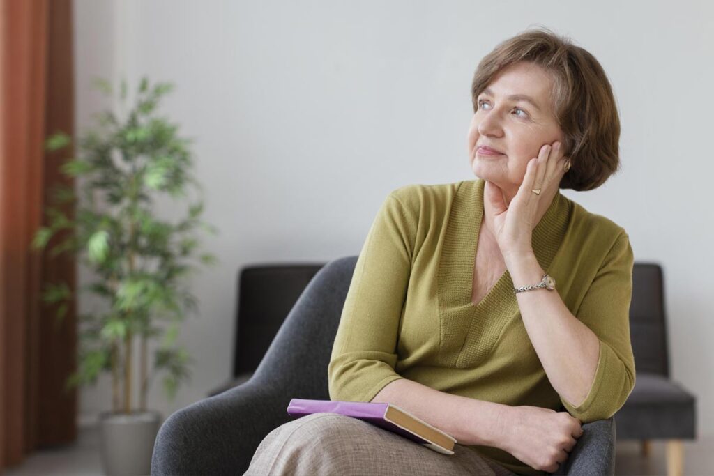 Femme d'une soixantaine d'années, vêtue d'un pull vert et d'un pantalon beige, assise dans un fauteuil chez elle, un livre reposant sur ses genoux. Elle semble absorbée par ses pensées.