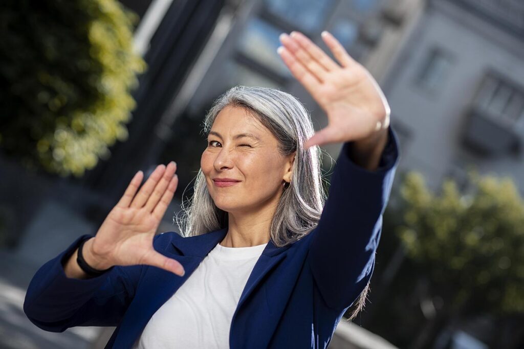 Femme dans la cinquantaine utilisant ses mains pour mimer le cadrage d'une photo, symbolisant son regard optimiste vers l'avenir et la confiance en soi.