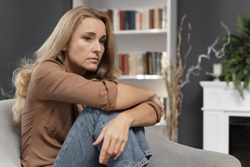 Femme symbolisant le manque de confiance en soi dans la quarantaine, blonde aux cheveux longs, vêtue d'un haut beige et d'un jean, apparaît triste et déprimée. Elle est assise sur son canapé, recroquevillée sur elle-même, les genoux remontés contre sa poitrine et enlacés par ses bras.