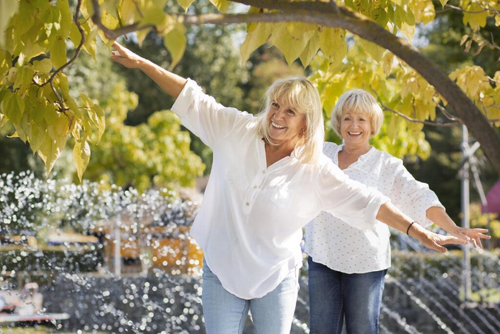 Deux femmes dans la soixantaine, reflétant une belle confiance en elles, toutes souriantes, vêtues de chemises blanches et de jeans, se promènent dans un parc orné d'arbres et de jets d'eau. Elles marchent l'une derrière l'autre, les bras étendus horizontalement pour garder l'équilibre.