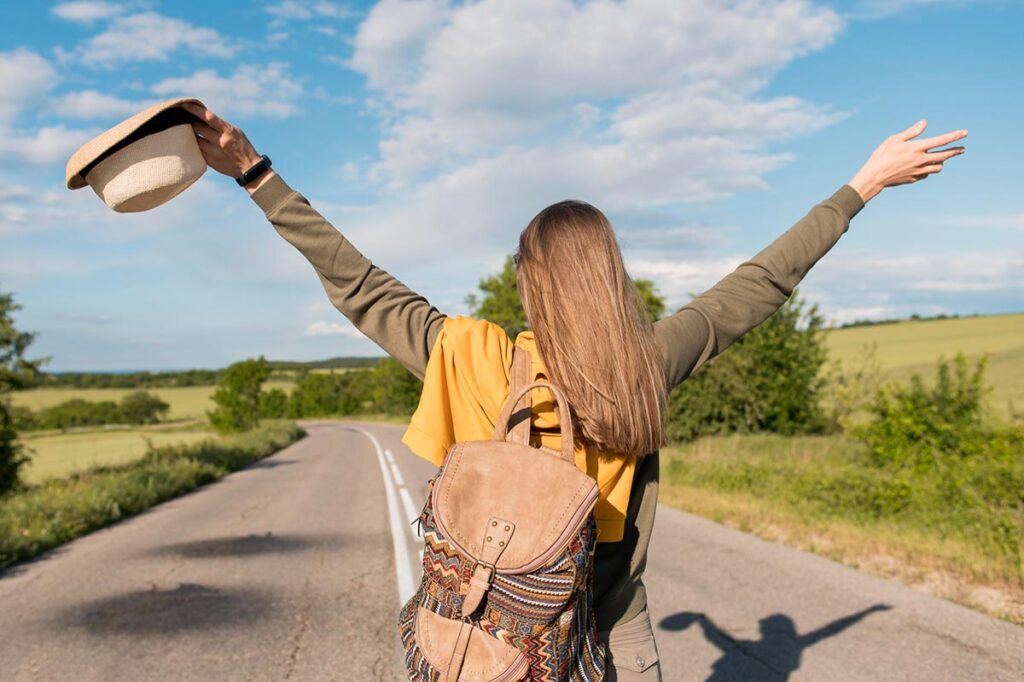 Une jeune femme aux longs cheveux blond châtain, vue de dos, lève les bras au ciel en tenant un chapeau de paille dans une main. Elle porte un haut et un pantalon beige, avec un pull jaune sur les épaules et un sac à dos. Elle se tient sur une route de campagne qui s'étend au loin. Cette image souligne les bienfaits de la marche à pied, mettant en avant la liberté et la joie que l'on peut ressentir lors d'une promenade à la campagne.