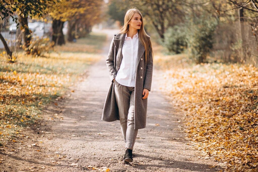 Une jeune femme élégante d'une quarantaine d'années marche d'un pas tranquille sur un sentier bordé d'arbres en automne. Elle porte un pantalon beige, un chemisier blanc et un long manteau ouvert beige, soulignant son style raffiné. Le sol est couvert de nombreuses feuilles mortes, ajoutant une ambiance saisonnière à la scène.