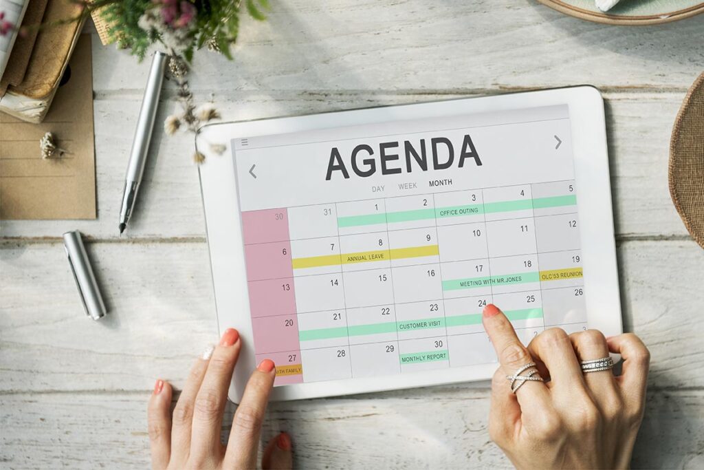 Gros plan sur un agenda posé sur une table en bois, avec un stylo à côté et des fleurs dans un pot. Les mains d'une femme avec du vernis à ongles rouge orangé pointent une date sur l'agenda. Cette image montre l'importance de bien choisir et planifier le moment de ses marches