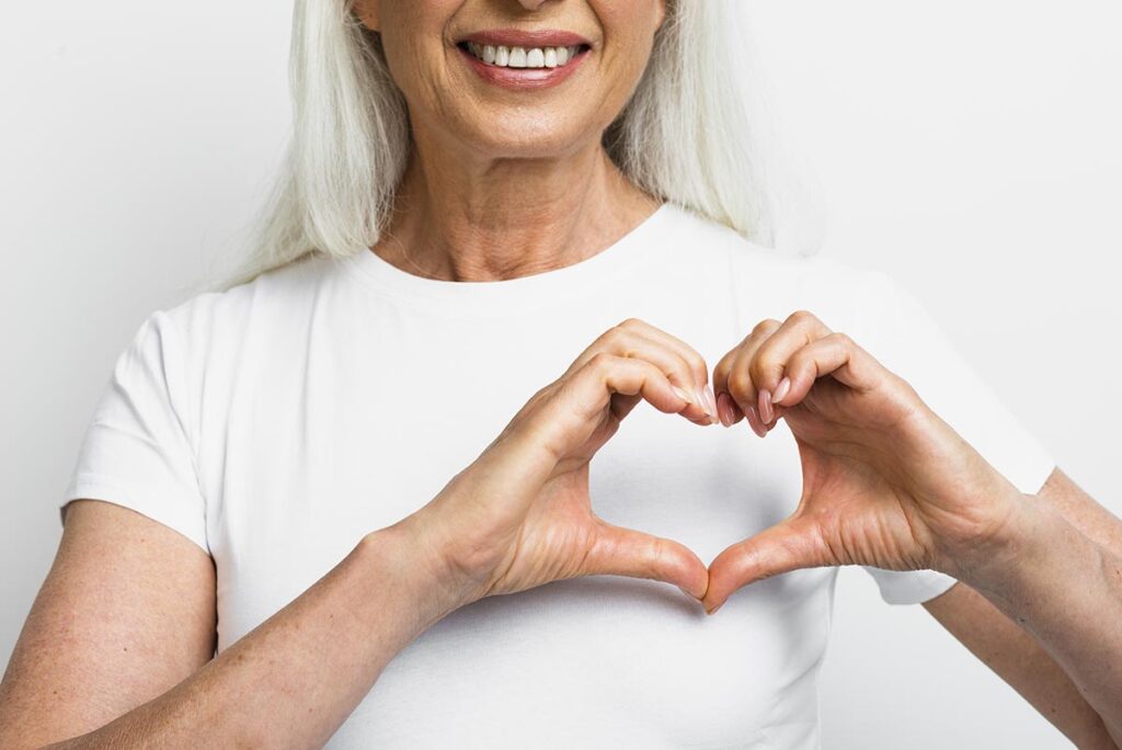 Une femme blonde en tee-shirt blanc, dont le haut du visage est hors-champ, forme un cœur avec ses doigts, symbolisant les bienfaits de la marche à pied.