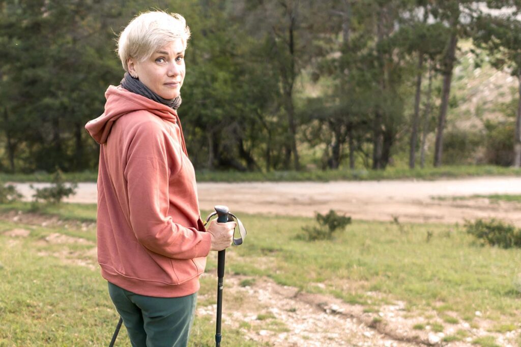 Une femme blonde aux cheveux courts, portant un pantalon vert et un sweat orangé, se tient à la lisière d'une forêt de pins. Elle est équipée de bâtons de marche, indiquant qu'elle pratique la marche à pied.