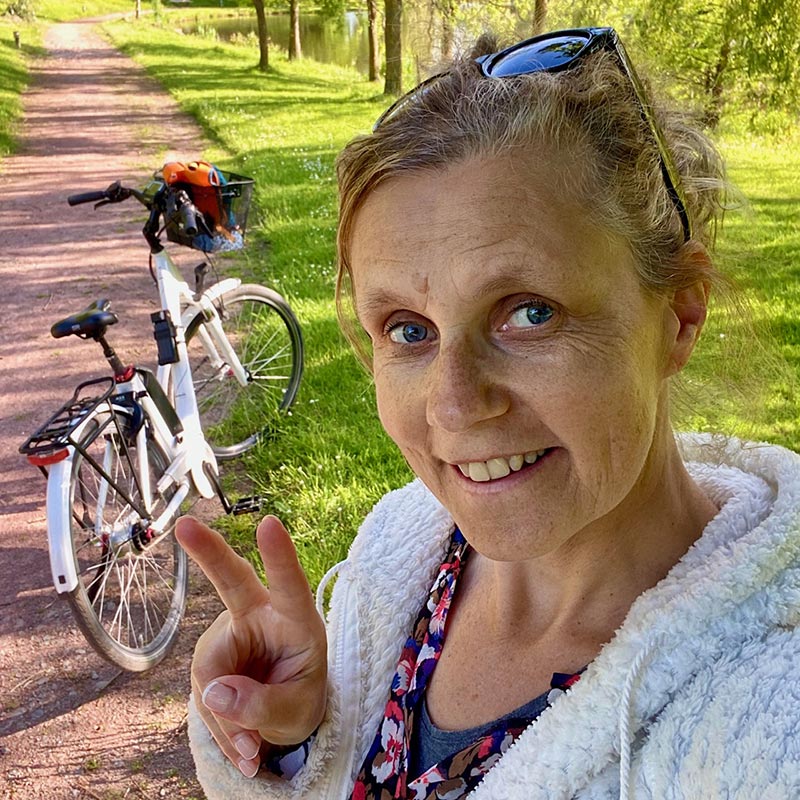 Moi, Béa, la fondatrice de Femmes en Harmonie, souriante et heureuse de revenir du travail à vélo, mes lunettes de soleil sur le haut du crâne, tenant mes cheveux. Je me suis débout sur un chemin rural avec en arrière-plan, mon vélo électrique sur sa béquille et mes affaires dans un panier accroché au guidon.
