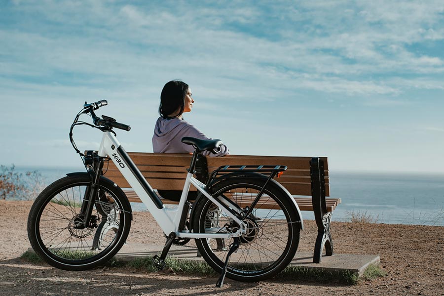 Une jeune femme brune assise sur un banc public face à la mer, avec son vélo électrique stationné derrière elle. Le ciel est bleu.