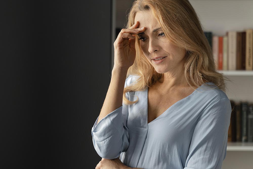 Une jeune femme d'une quarantaine d'années, blonde aux cheveux longs, est vêtue d'un chemisier bleu ciel. Elle se tient la tête, visiblement préoccupée. Son expression trahit des pensées tournées vers le passé, regrettant ce qu'elle a perdu ou manqué. Cette fixation l'empêche de vivre pleinement le présent.
