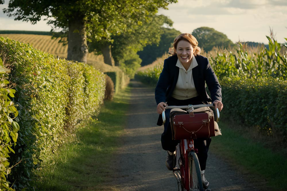 Une jeune femme rousse en costume de ville pédalant à vive allure sur un chemin de campagne bordé d'arbres, de verdure et d'un champ de maïs.