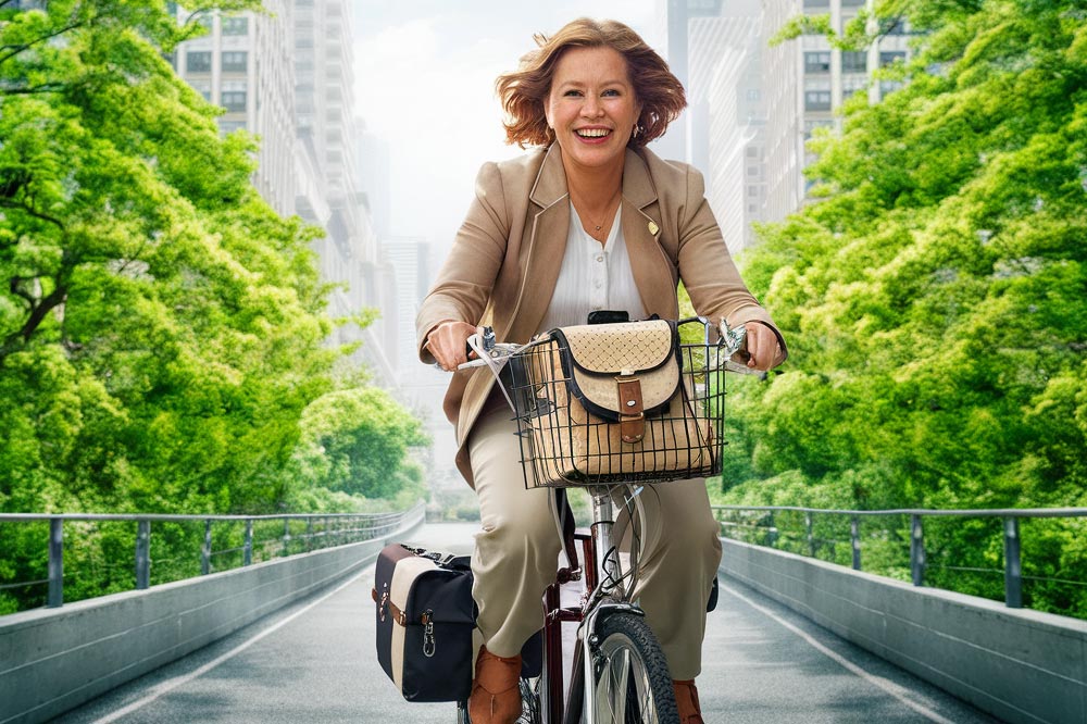 Une femme entre cinquante et soixante ans, les cheveux au vent et souriante, pédale sur une piste cyclable. Son sac à main est dans un panier accroché au guidon et son vélo est équipé de sacoches sur le porte-bagages.