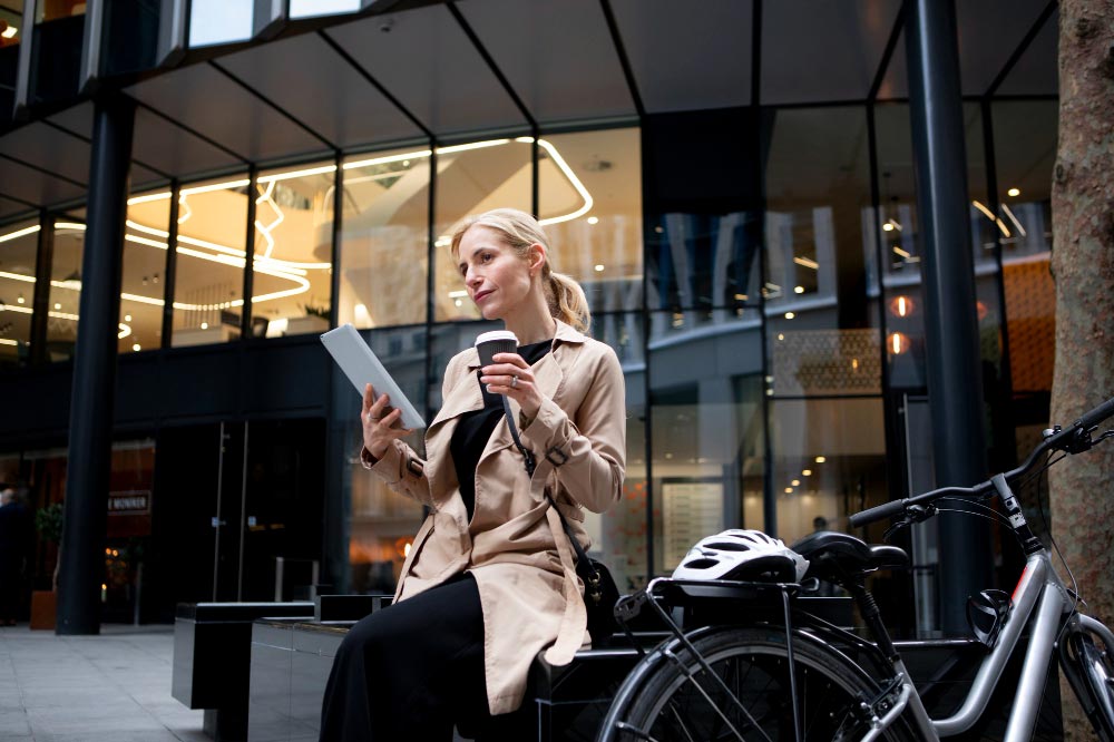 Une femme blonde aux cheveux longs attachés en queue de cheval, portant un imperméable crème, lit sa tablette et apprécie un café. Elle est assise sur un mobilier urbain avec un grand sourire, heureuse avec son deux-roues à côté d'elle, sur lequel est posé son casque.