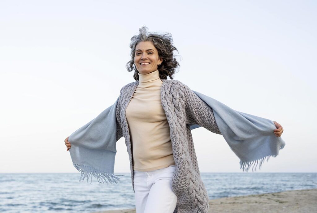 Sur la plage, une femme entre cinquante et soixante ans court avec un immense sourire, les bras tendus à l'horizontal telles des ailes, exprimant pleinement la joie de vivre. Vêtue d'un sous-pull jaune, d'un long gilet en laine gris et d'un châle bleu ciel qu'elle porte sur ses bras, elle incarne la liberté et l'épanouissement. Cette image captivante nous rappelle l'importance de savourer chaque instant, de se laisser porter par la légèreté du moment présent et de cultiver une joie authentique au quotidien.