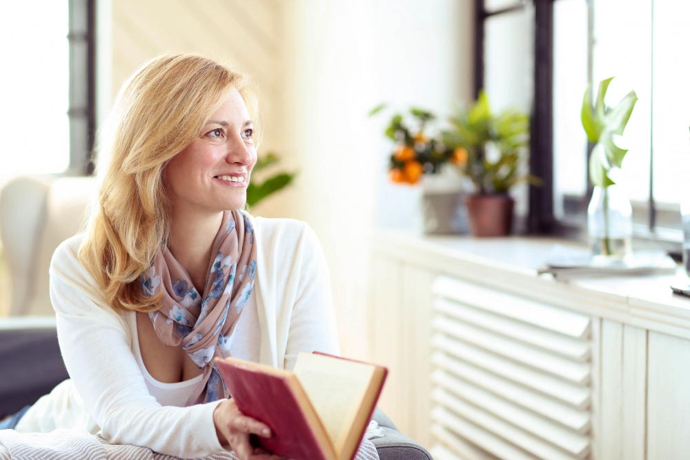 Une femme blonde aux cheveux longs, dans la quarantaine, porte un gilet blanc et un foulard multicolore, assise dans son salon, un livre entre ouvert entre ses mains. Elle sourit en regardant par la fenêtre, se projetant vers son avenir tout en appréciant l'instant présent.