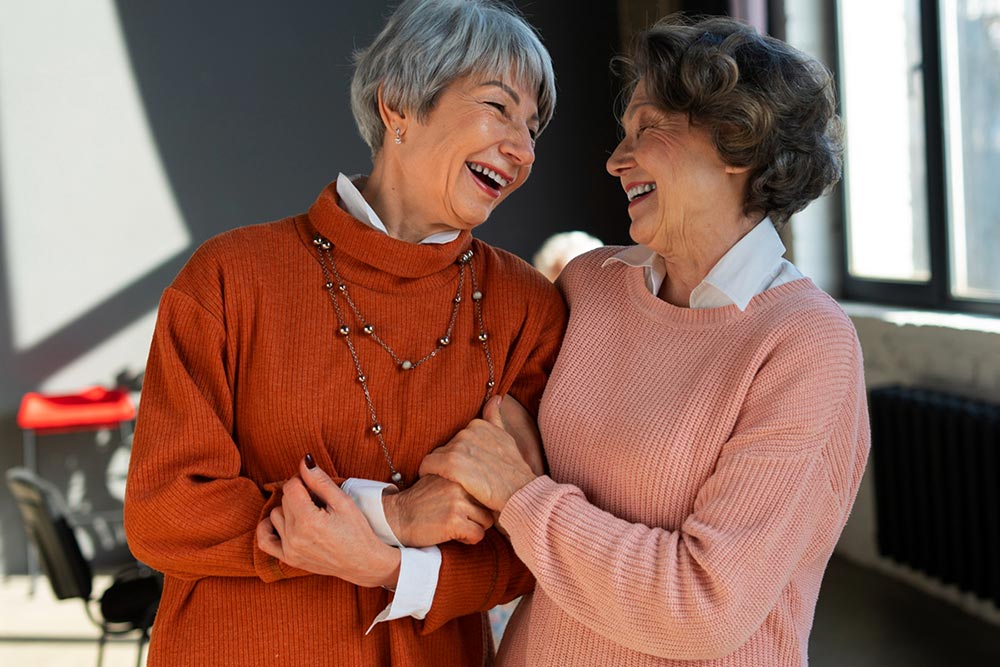 Deux amies d'environ cinquante ou soixante ans partagent un éclat de rire en se tenant les bras. Celle de gauche, aux cheveux courts et gris, porte un pull orange laissant entrevoir le col d'un chemisier blanc, agrémenté d'un grand collier composé de boules métalliques. La seconde, à droite, aux cheveux mi-longs et bruns, arbore un pull rose dont dépasse également le col d'un chemisier blanc. Ce moment de complicité illustre une amitié solide et rappelle qu'il y a de la joie dans les petites choses, que ce soit un repas partagé avec des amis, une marche à pied dans la nature, ou simplement la lecture d’un bon livre.