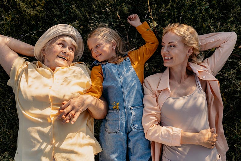 Une grand-mère d'environ soixante-dix ans, vêtue d'une robe jaune et coiffée d'un chapeau de paille, est allongée dans l'herbe d'un jardin. À ses côtés, sa petite-fille d'environ 8 ou 10 ans, aux cheveux blonds longs, porte une salopette en jean et un sous-pull orange. À côté d'elles, sa fille d'environ 45 ans, aux cheveux blonds/roux, vêtue d'un haut beige et d'une veste légère rose, partage un moment de complicité. Les trois affichent un grand sourire, exprimant la joie et le bonheur du moment présent passé ensemble.