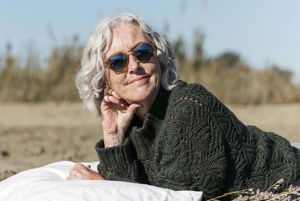 Une femme entre soixante et soixante-dix ans, allongée sur le ventre sur un oreiller, rayonne de bonheur. Avec ses longs cheveux gris, ses lunettes de soleil et son pull en laine vert foncé, elle vit pleinement sa vie dans l'harmonie et l'épanouissement. Son sourire témoigne de sa joie authentique et de sa sérénité intérieure.