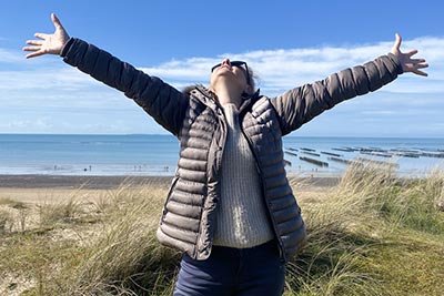 Fondatrice de Femmes en Harmonie, Béa se tient dans les dunes avec la mer en arrière-plan, les bras levés vers le ciel et un grand sourire. Elle montre le chemin aux femmes de la communauté, les encourageant à cultiver une attitude positive et à vivre pleinement chaque instant.