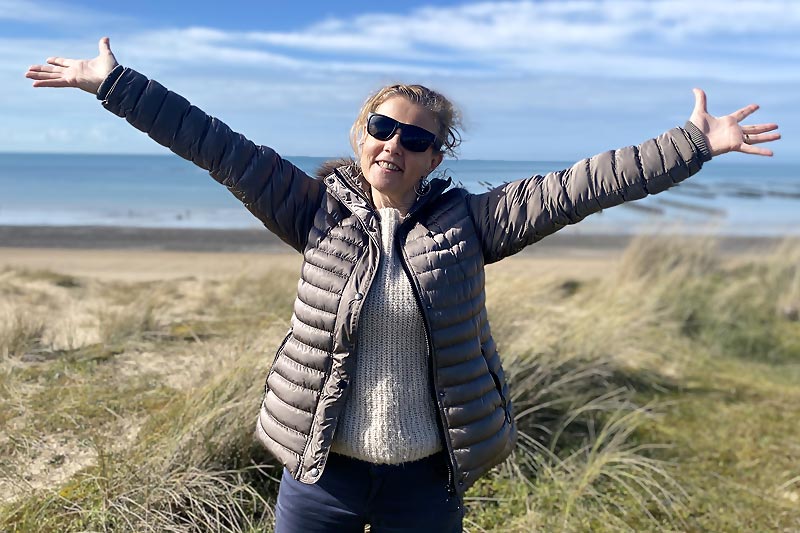 Béa, créatrice de Femmes en Harmonie, irradie de joie parmi les dunes avec l'océan en toile de fond. Arborant un large sourire, elle lève les bras vers le ciel, illustrant l'importance de cultiver une attitude positive et de vivre pleinement avec reconnaissance.
