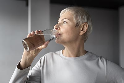 Une femme de soixante ans, aux cheveux gris et courts, boit de l'eau à la bouteille. La déshydratation peut affecter ton niveau d'énergie et ta capacité de concentration.