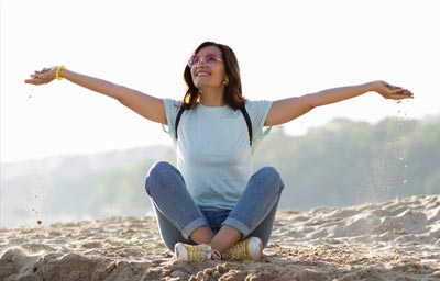 Une femme brune aux cheveux longs, avec un grand sourire, est assise en tailleur sur une plage, les bras grands ouverts, illustrant parfaitement comment cultiver une attitude positive peut enrichir la vie. Sa posture ouverte et son expression joyeuse capturent l'essence du bien-être émotionnel et de la pleine conscience