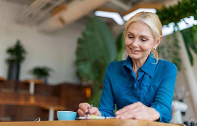 Une femme d'environ 60 ans, les cheveux blonds attachés en queue de cheval et portant un chemisier bleu, rédige son journal de gratitude assise à une table. Elle incarne l'importance de cultiver une attitude positive en prenant le temps de reconnaître et d'apprécier les aspects positifs de sa vie.