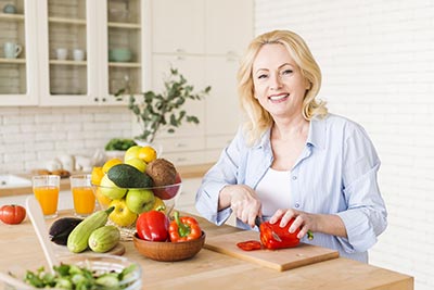 Dans sa cuisine, une femme blonde aux cheveux longs, portant un chemisier bleu ciel ouvert sur un tee-shirt blanc, coupe différents légumes sur son plan de travail. Une bonne alimentation fournit les nutriments nécessaires pour soutenir ton bien-être physique et mental.