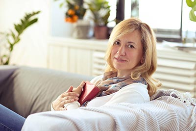Une femme élégante de 50 ans, blonde aux cheveux longs, se repose assise confortablement dans son canapé, tenant un livre posé sur sa poitrine. Prendre le temps de lire et de se détendre aide à cultiver une attitude positive et à nourrir son esprit.