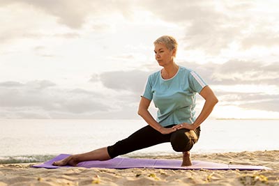 Une femme d'environ soixante ans, en tenue de sport, fait des étirements sur une plage. Elle illustre l'importance de cultiver une attitude positive en prenant soin de son corps et en appréciant l'exercice en plein air.
