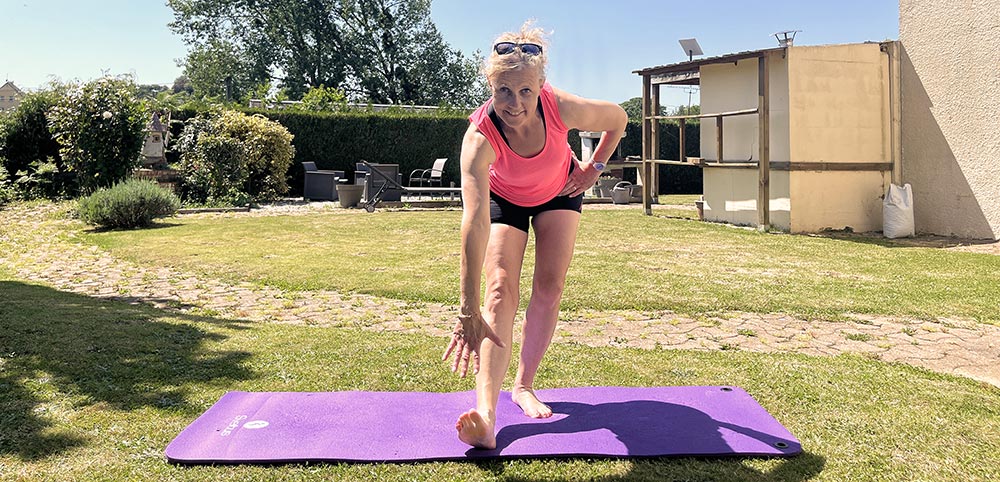 Sur un tapis de sport posé sur la pelouse de son jardin, Béa, la fondatrice de Femme en Harmonie, sourit en montrant comment réaliser techniquement et correctement des exercices étirements.