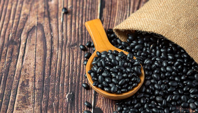 Sac de haricots noirs sur une table en bois vernis avec une cuillère remplie de haricots sortant du sacs.