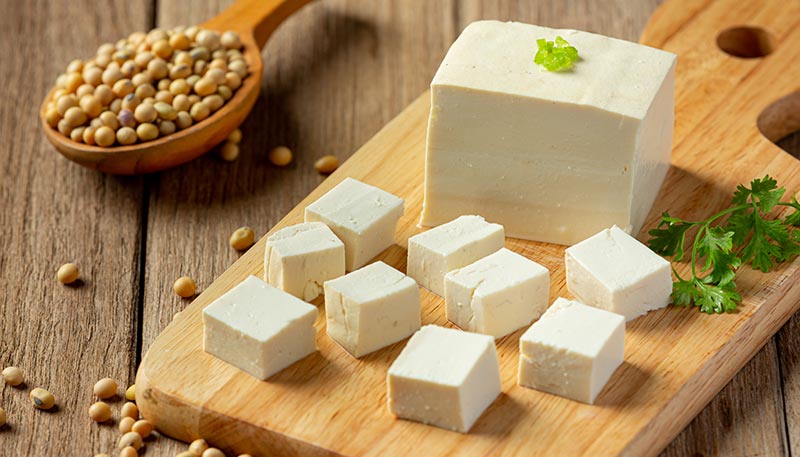 Gros cube de tofu à côté de plusieurs petits cubes fraîchement découpés sur une planche en bois posée sur une table rustique.