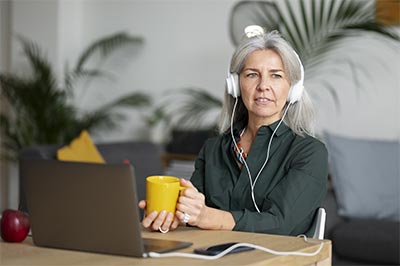 Femme d'une cinquantaine d'années assise à une table chez elle, devant son ordinateur portable, tenant une tasse jaune entre les mains et portant un casque audio, illustrant l'importance du conditionnement mental positif le matin.
