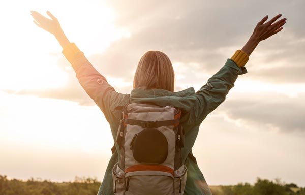 Femme blonde aux cheveux longs de dos, portant une veste verte et un sac à dos, les bras levés vers le ciel au petit matin, symbolisant l'intégration des affirmations positives dans la routine quotidienne.