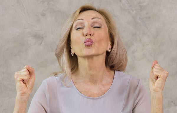 Femme d'environ soixante ans, blonde aux cheveux longs, portant un pull mauve, les yeux fermés et les poings serrés, illustrant le renforcement de la confiance en soi avec les affirmations positives du matin.