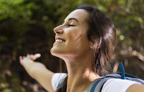 Femme brune aux cheveux longs attachés en arrière, les yeux fermés et les bras ouverts, souriant dans la nature, illustrant l'importance de combiner affirmations positives et exercices de respiration pour une synergie corps-esprit