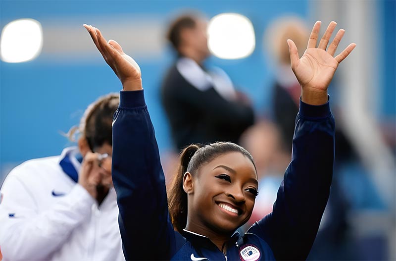 Simone Biles, la gymnaste américaine légendaire, avec un grand sourire et les bras levés pour saluer le public après une épreuve. Cette photo capte le moment de célébration de Simone Biles, illustrant sa joie et son triomphe. Elle incarne non seulement l'excellence dans la gymnastique mais aussi la résilience et la détermination, faisant d'elle un modèle de force intérieure et d'inspiration dans le monde du sport.