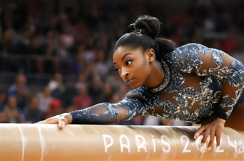 La gymnaste américaine Simone Biles, concentrée et allongée sur la poutre des JO 2024 à Paris, où est inscrit 'PARIS 2024', montre une maîtrise technique exceptionnelle. Cette photo capture son retour triomphal lors de la compétition, illustrant son niveau de performance remarquable.