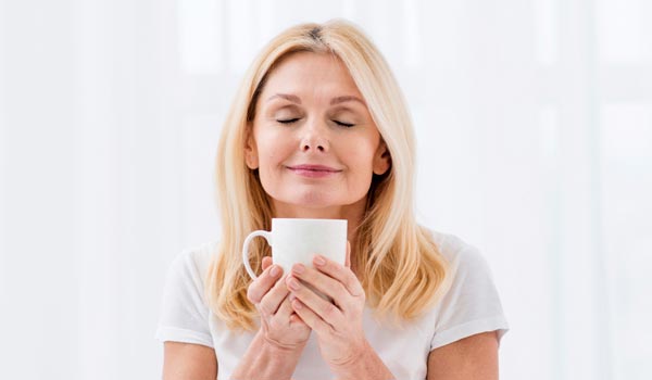 Femme blonde de 50 à 55 ans, souriante, tenant une infusion dans ses mains, symbole de détente et de bien-être. Elle incarne l'idée que des choix alimentaires réguliers et sains, comme les aliments anti-inflammatoires, peuvent aider à réduire l'inflammation dans le corps et renforcer l'immunité.
