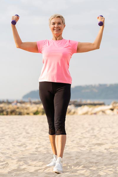 Femme blonde d'une cinquantaine d'années, levant de petites haltères sur la plage, illustrant l'importance d'intégrer l'exercice physique dans la routine quotidienne.