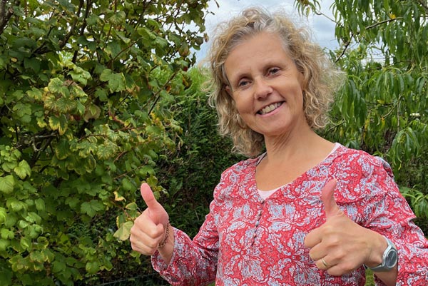 Béa, la fondatrice de Femmes en Harmonie, très souriante faisant un pouce levé dans son jardin. Elle illustre la joie de vivre et l'engagement envers une mentalité positive.